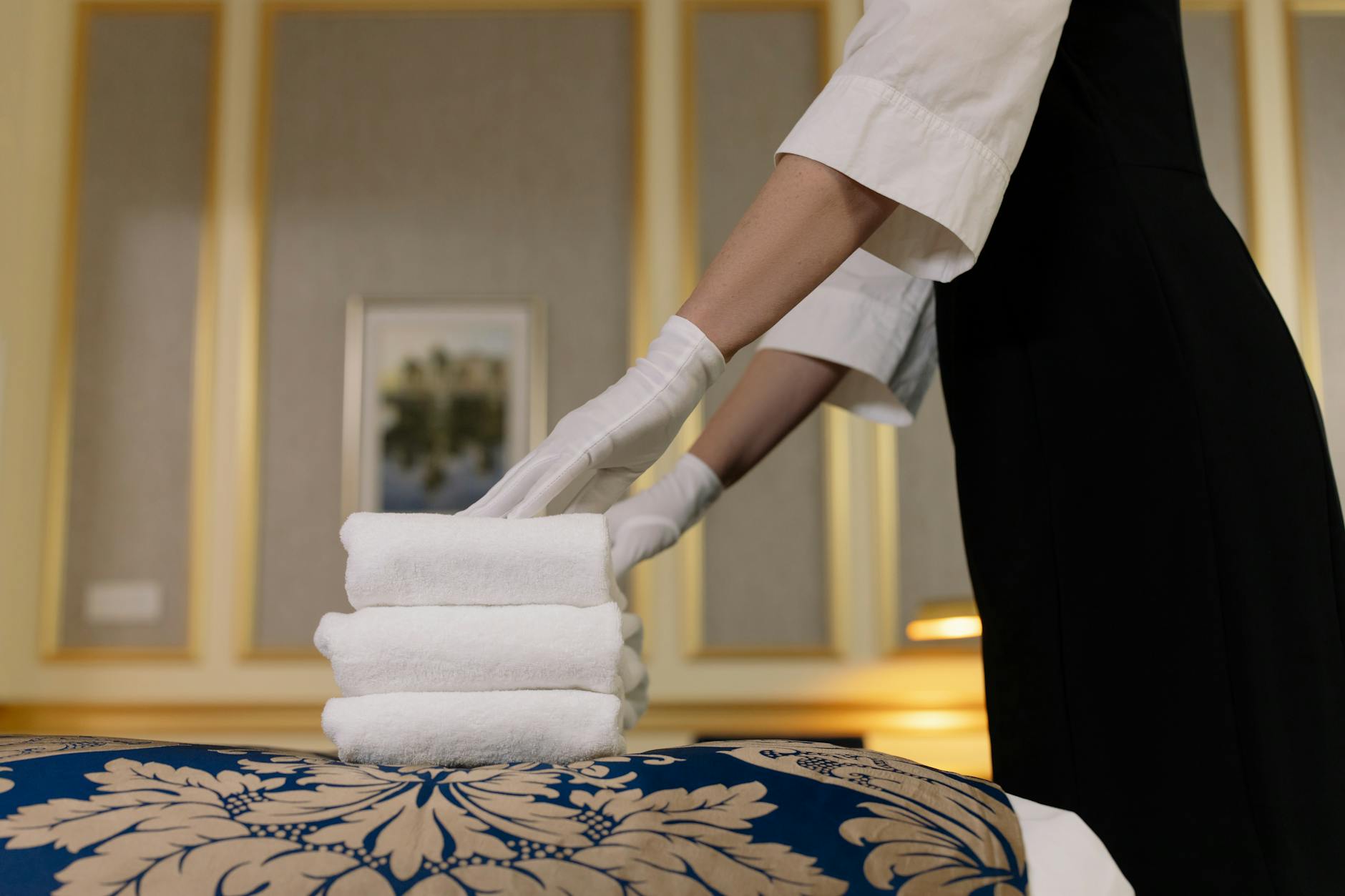 Hotel staff member wearing white gloves neatly folding and stacking fluffy white towels on a freshly made bed, showing attention to cleanliness and detail.