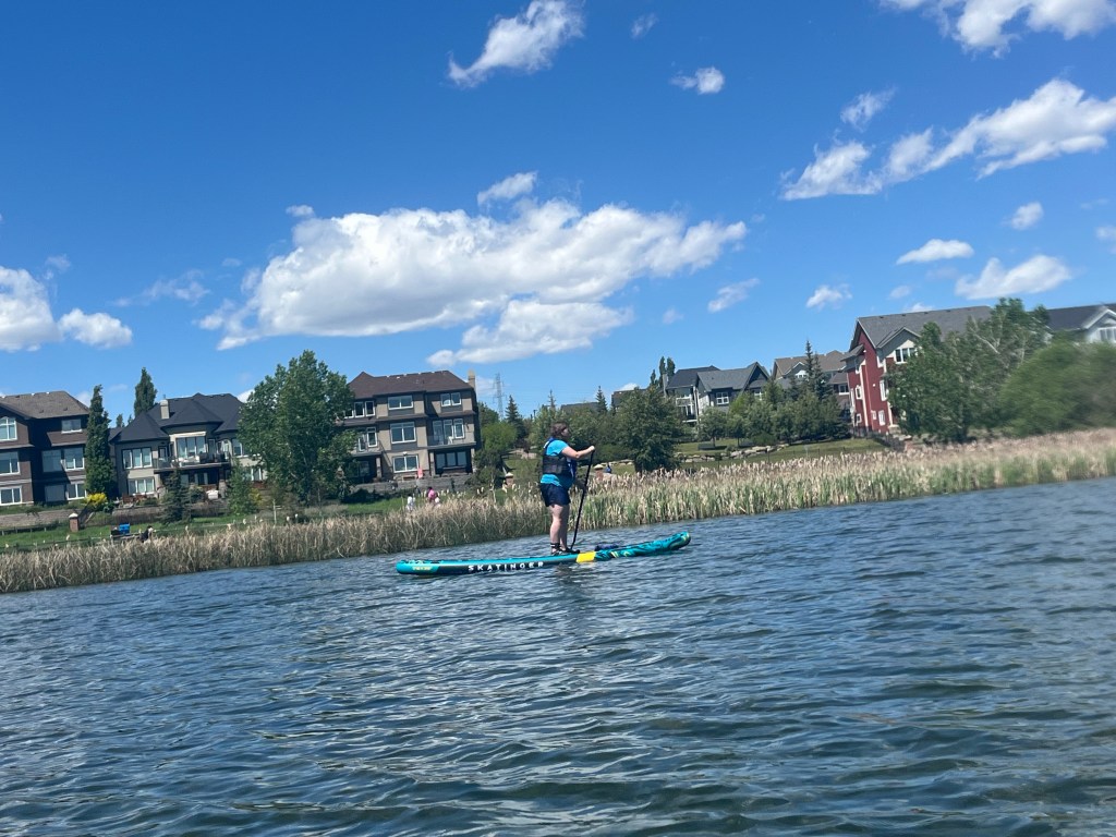 Christine standing on a paddle board at Chestermere lake. With houses in the background and a blue sky. 