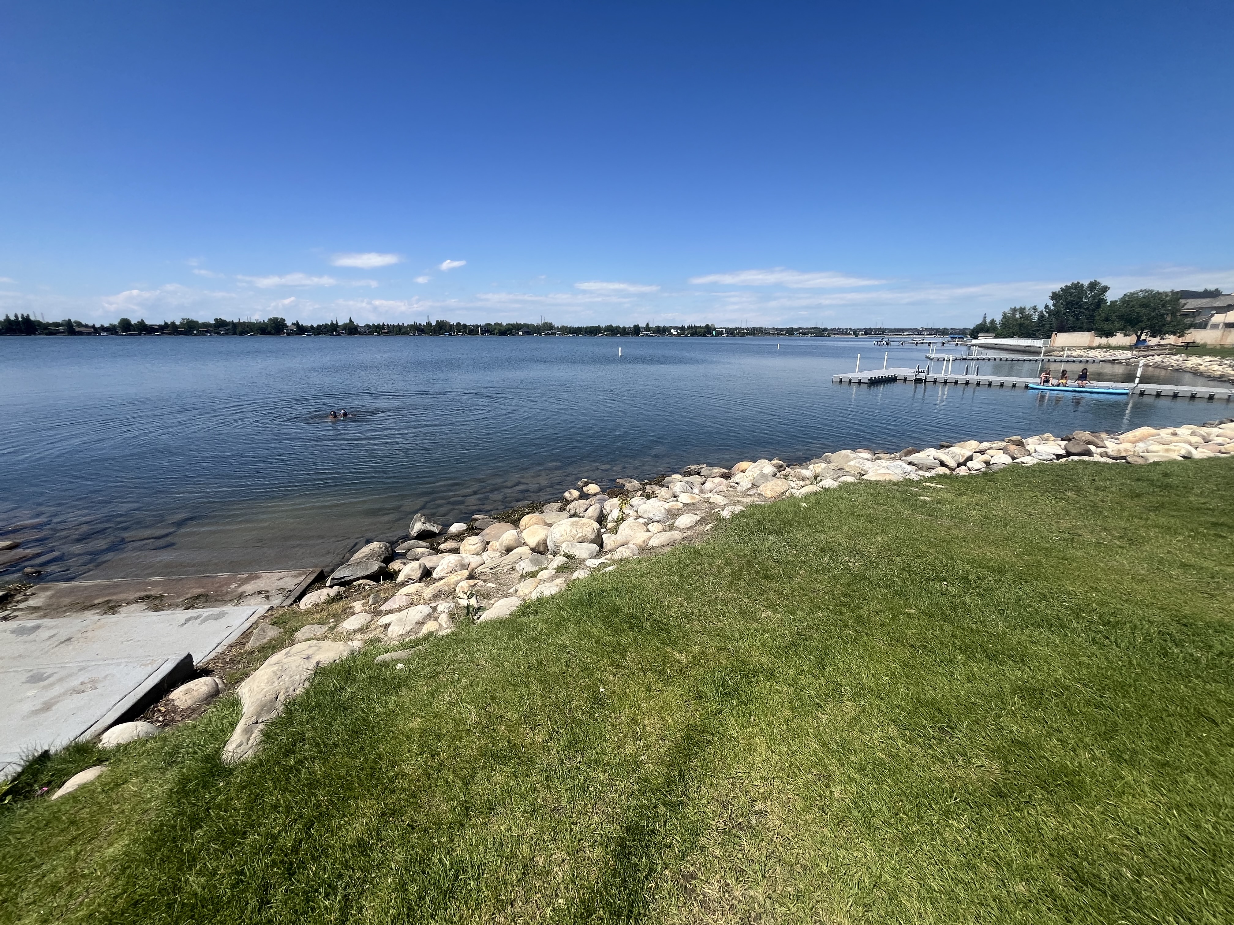 Blue Sky, blue crisp water, rocky shore line that breaks with stairs leading up to the grassy area of people to relax and play in. In the distance you can see two floating docks. 